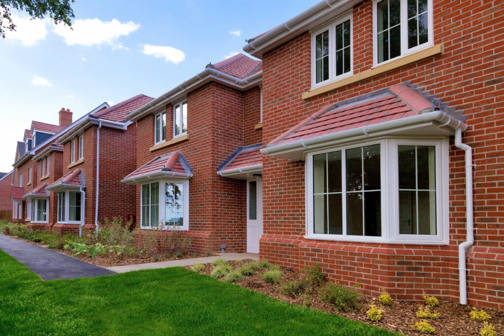 Row of modern red-brick houses with large bay windows, white frames, and manicured front lawns, under a blue sky with light clouds. Shrubs and a paved path run alongside the homes.