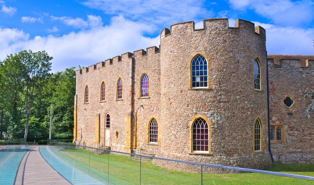 A historic stone castle with arched windows and crenellated battlements stands beside a modern glass walkway, surrounded by grass and trees under a bright blue sky with scattered clouds.