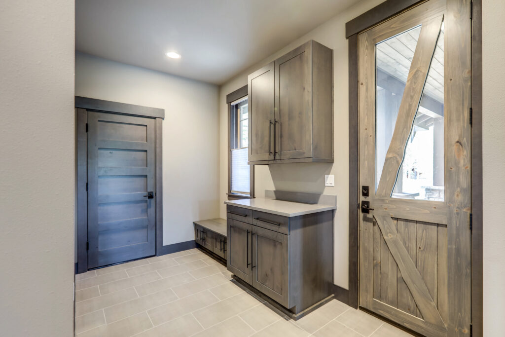 Mudroom with light tiled floor, rustic wooden cabinets, a bench with storage, a window, and two wooden doors—one interior, one exterior with a diagonal cross brace and glass panel. Neutral color palette and recessed lighting.
