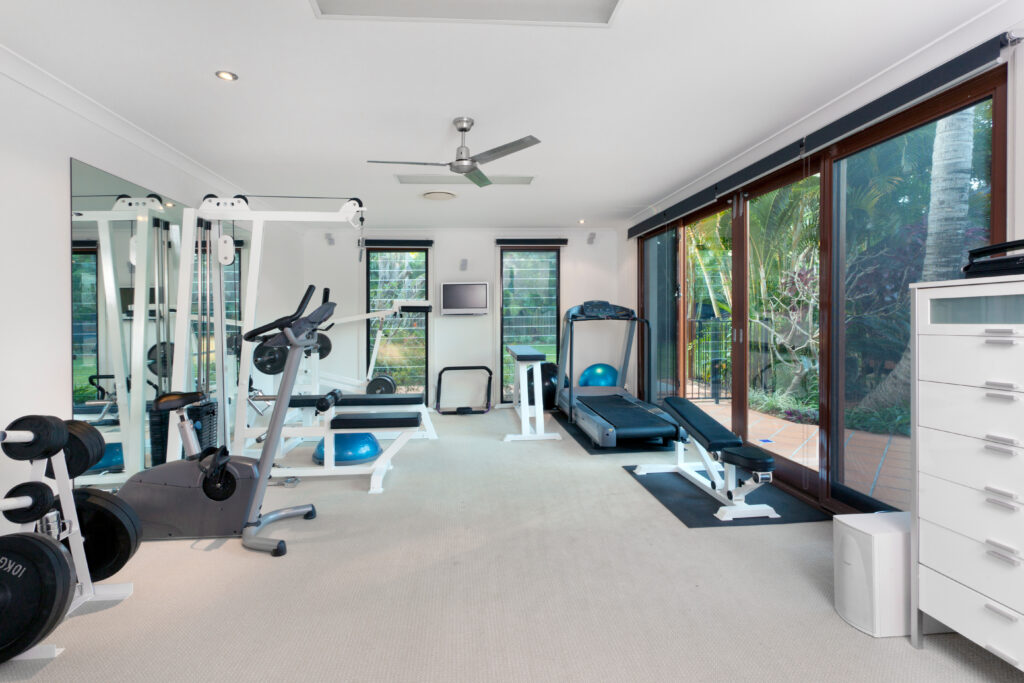 A bright home gym with exercise equipment, including a treadmill, stationary bike, weight machine, dumbbells, and yoga balls, next to large windows overlooking a garden.