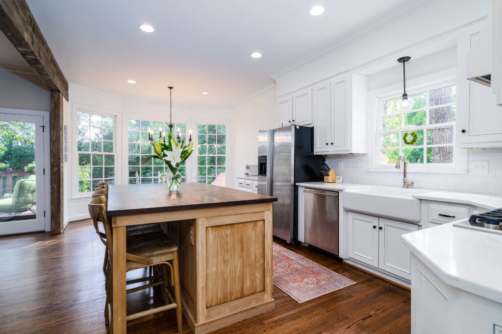 Bright kitchen with white cabinets, stainless steel appliances, a large wooden island with chairs, farmhouse sink, hardwood floors, flowers on the island, and large windows letting in natural light.