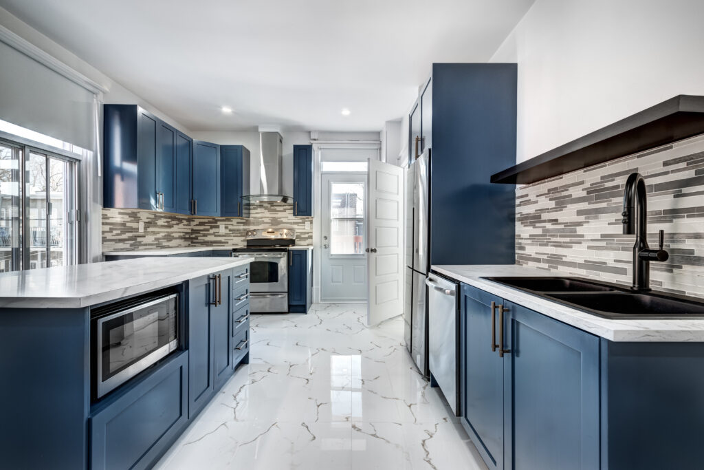 Modern kitchen with blue cabinets, white marble floors, stainless steel appliances, a black sink, and a gray-white tile backsplash. Large windows and a glass door let in natural light.