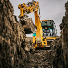 A person operates a yellow excavator, digging a deep trench in the earth. The photo is taken from inside the trench, showing soil walls on both sides and cloudy sky above.