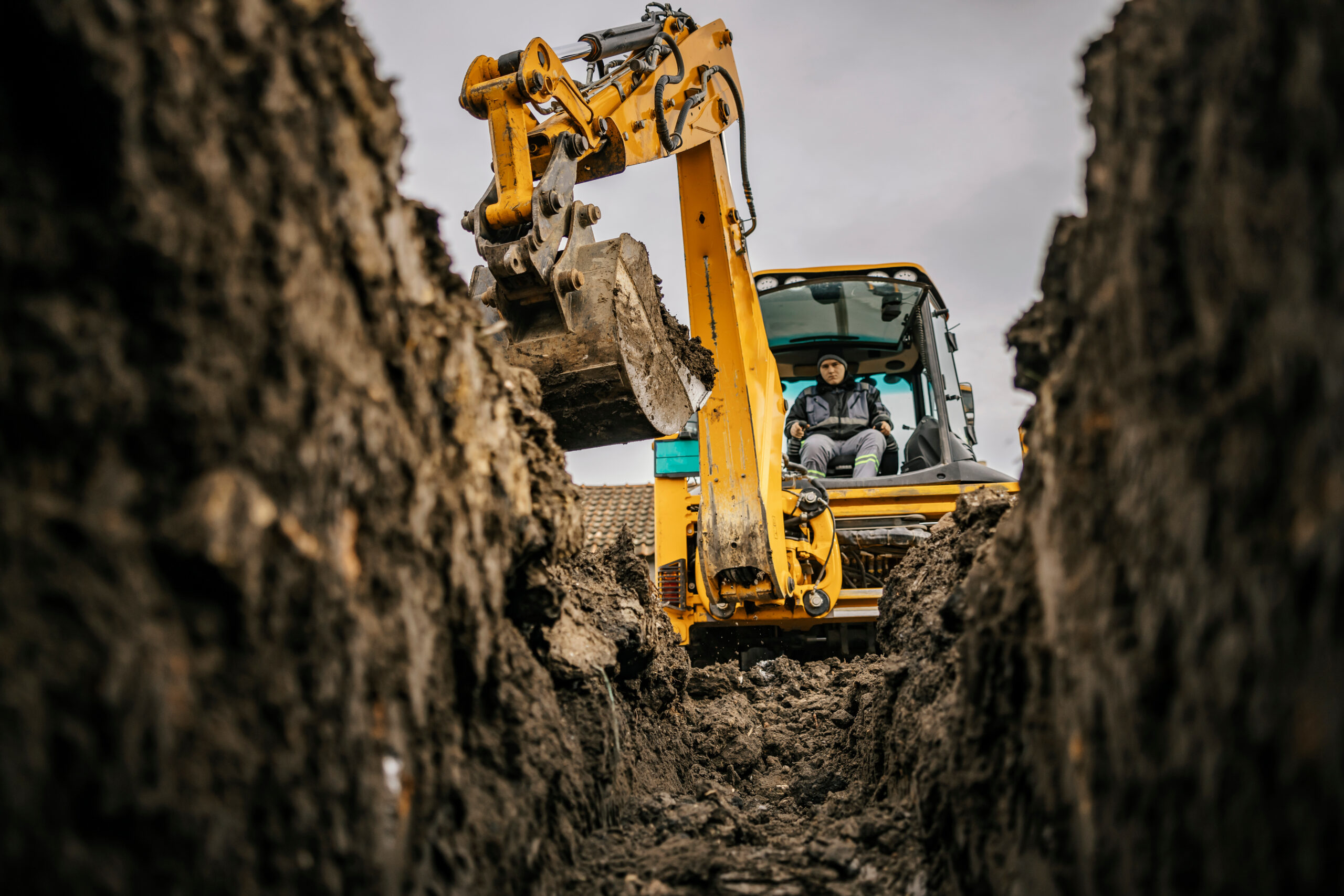 A person operates a yellow excavator, digging a deep trench in the earth. The photo is taken from inside the trench, showing soil walls on both sides and cloudy sky above.