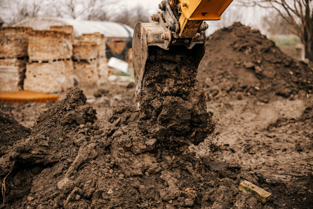 A close-up of an excavator’s bucket lifting a pile of dirt at a construction site, with scattered soil and blurred brick structures in the background.