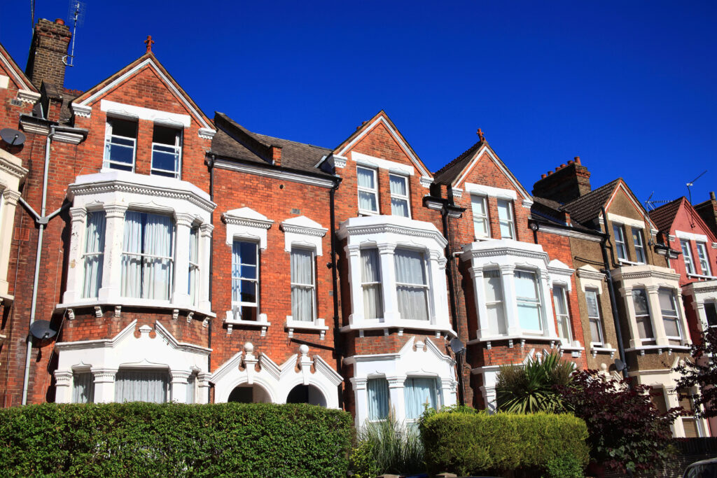 A row of Victorian-style red brick townhouses with white trim and large bay windows, set against a clear blue sky, with green hedges and shrubs in front.