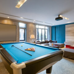 A modern lounge with a blue pool table set for a game, surrounded by sofas, armchairs, a coffee table, large windows, a blank screen on the wall, and ceiling lights creating a bright, inviting atmosphere.