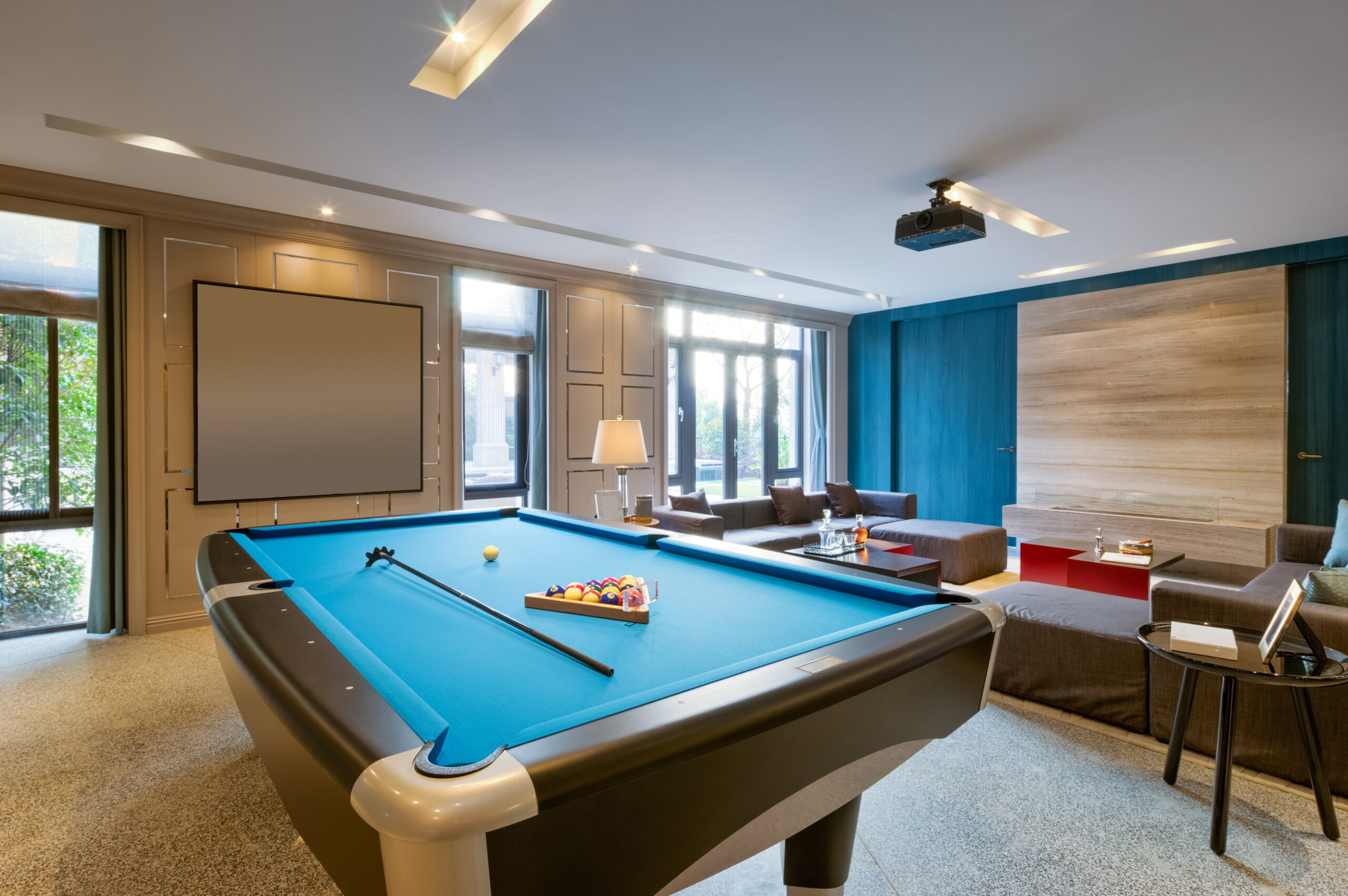 A modern lounge with a blue pool table set for a game, surrounded by sofas, armchairs, a coffee table, large windows, a blank screen on the wall, and ceiling lights creating a bright, inviting atmosphere.