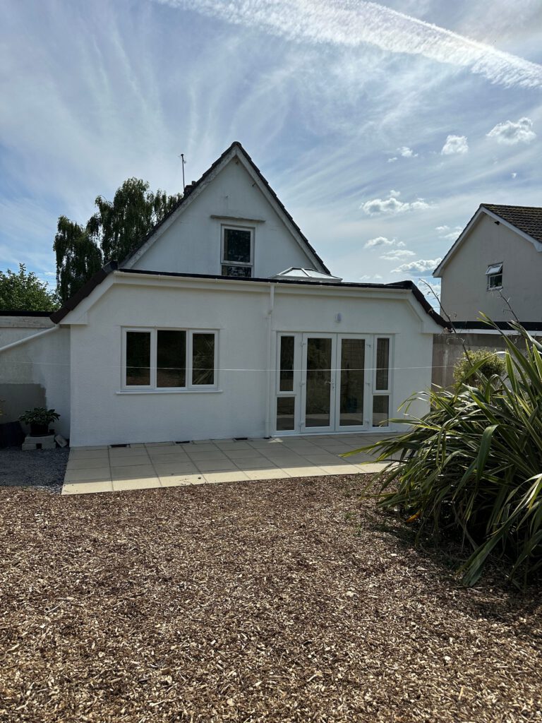 A modern white house with a sloped roof and large windows facing a patio with paving stones and a small mulched garden area, under a blue sky with wispy clouds.