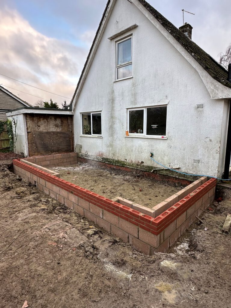 A white house with a partially built brick and block extension foundation in the foreground, surrounded by dirt and construction materials under a cloudy sky.