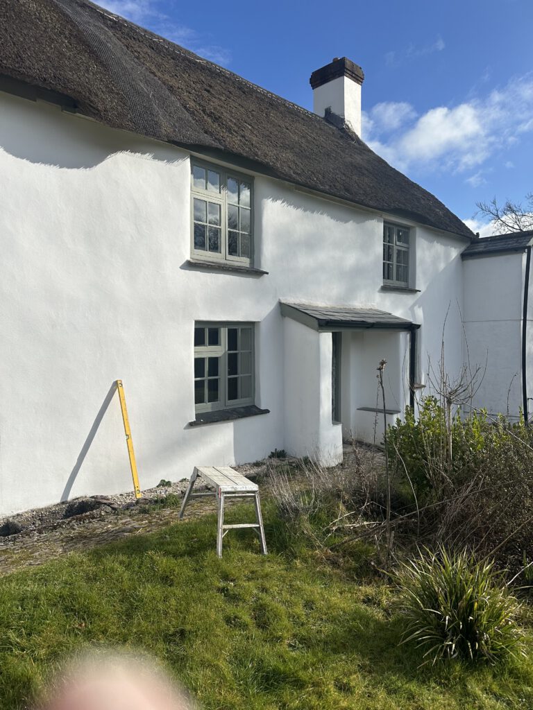 A white, two-story cottage with a thatched roof and gray-trimmed windows, set under a blue sky. A small stepladder, measuring stick, and overgrown grass are visible in the garden at the front.