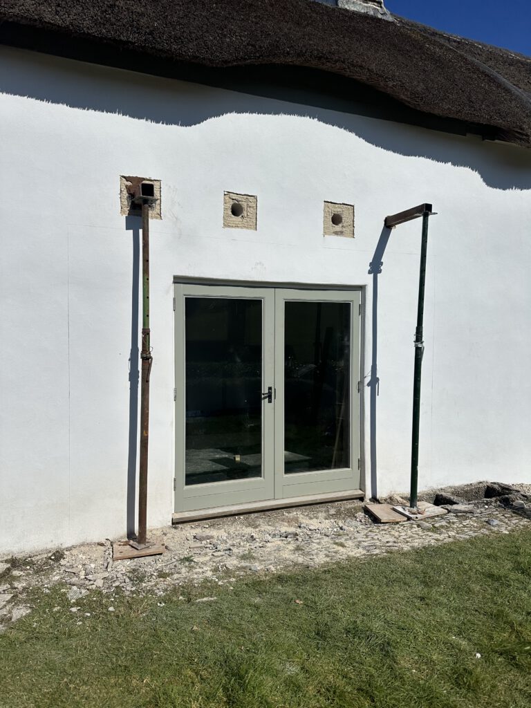 A pair of glass double doors set in a white wall, with three square holes above them and two metal support poles on either side. The ground in front is bare earth and patches of grass.