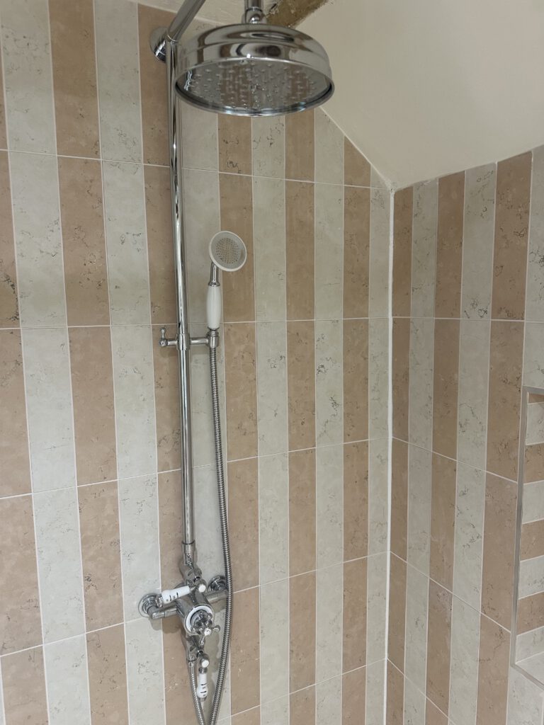 A walk-in shower with beige and cream vertical tiles, featuring a large round overhead showerhead and a smaller handheld shower attached to a vertical metal bar.